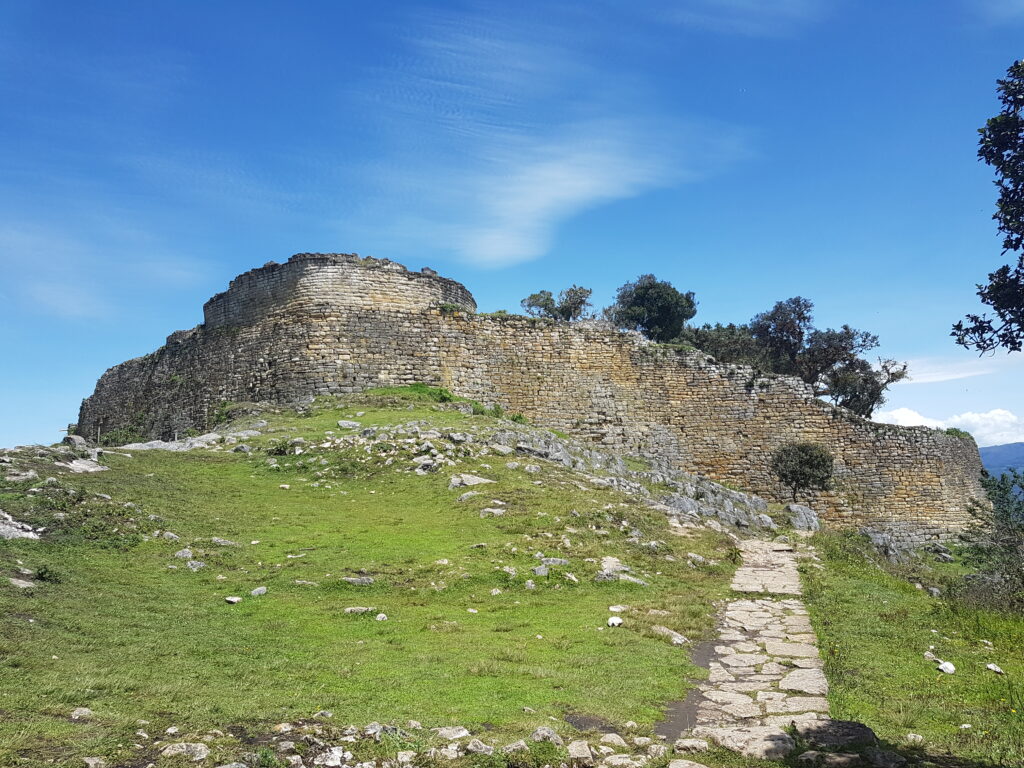 The outer wall of the Kuelap fortress under a blue sky, with a stone path leading towards it.