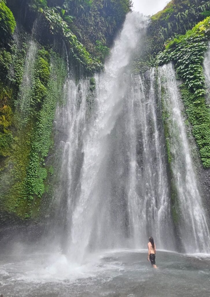 Person standing in the pool beneath Tiu Kelep waterfall in Lombok, surrounded by moss-covered cliffs.