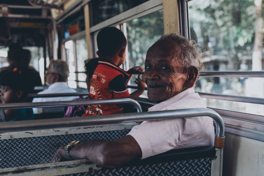 Elderly man in a white shirt smiling over his shoulder on a crowded local bus in Sri Lanka