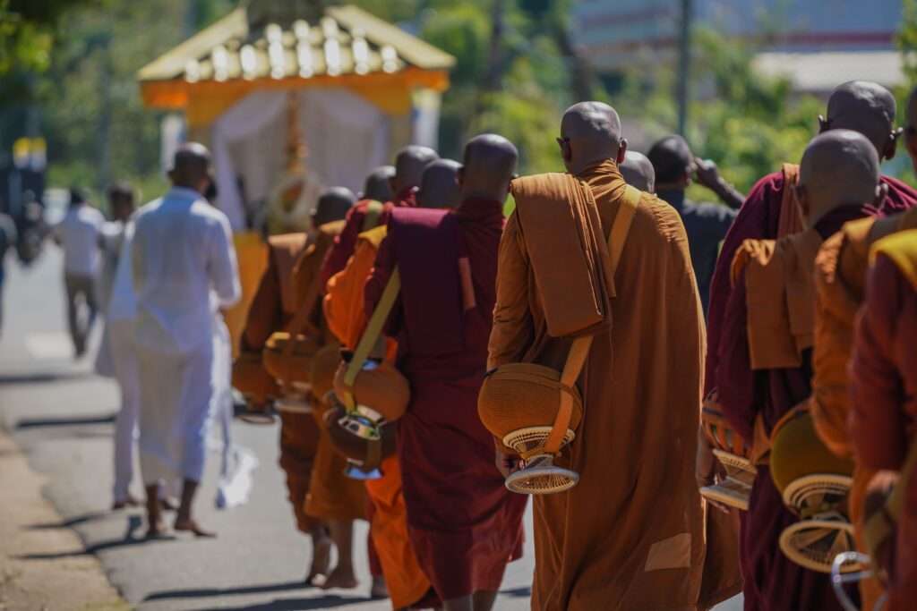 Buddhist monks in orange and maroon robes walking with alms bowls through a street in Sri Lanka